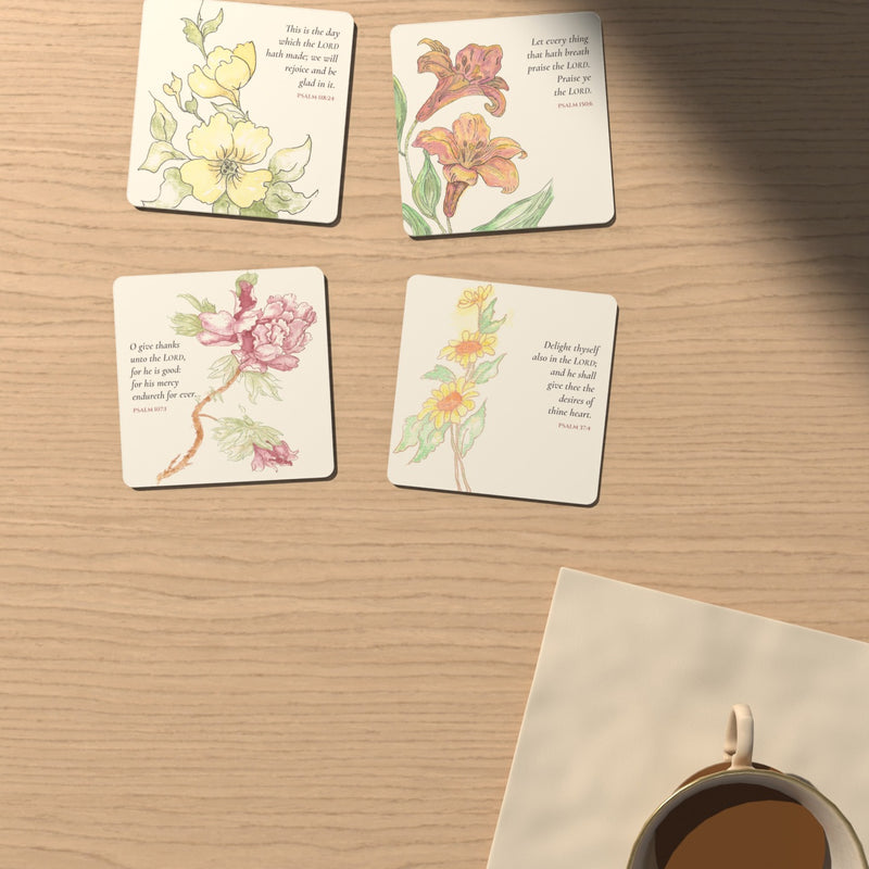 Four watercolor Psalm coasters on a light oak table beside a cup of tea: yellow blossoms, orange lilies, a pink peony and yellow daisies