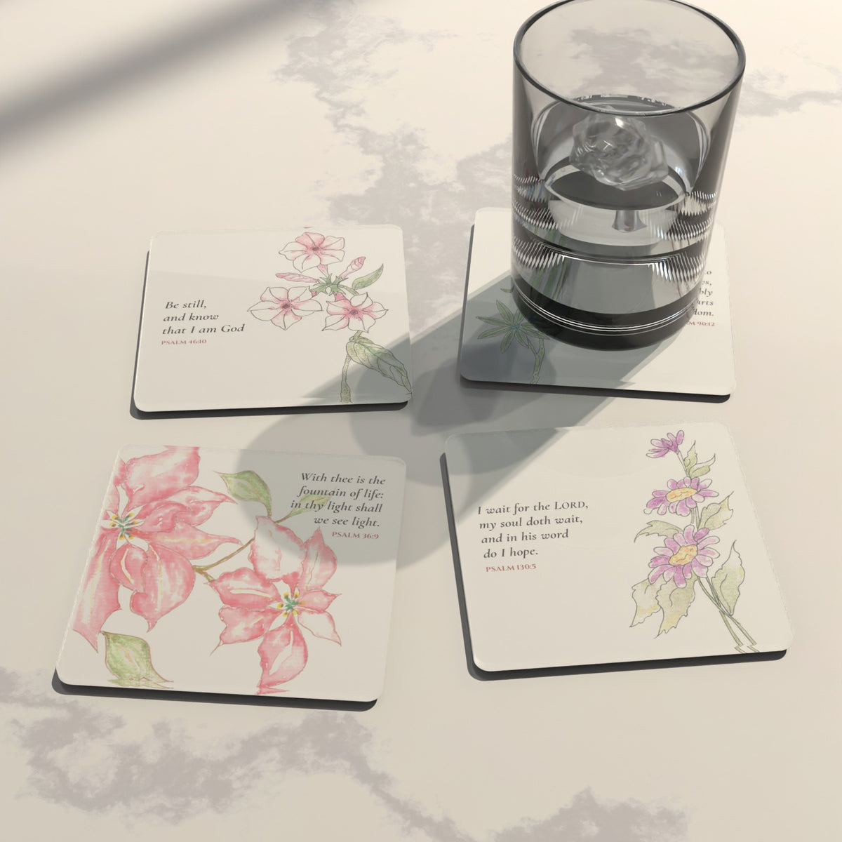 Four watercolor Psalm coasters on white marble with a glass of ice water: pink blooms, purple asters, pink phlox and bluebonnets